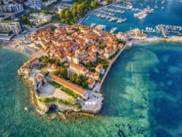 Veduta aerea della penisola del centro storico di Budva protesa nell'Adriatico turchese, con l'abitato compatto entro le mura e le spiagge circostanti