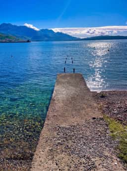A pier reaching into calm water at Bijela, with the pebble shoreline and bay beyond.