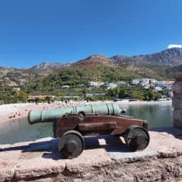 Cañón de bronce histórico en el muro de piedra de Sveti Stefan con la costa de Paštrovići y las montañas detrás