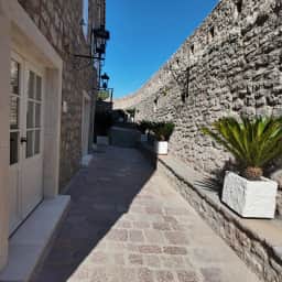 Una estrecha calle pavimentada entre una cabaña blanca y un alto muro de piedra dentro de Sveti Stefan