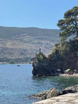 The ballerina statue seen from along the seaside footpath between Budva Old Town and Mogren beach, with walkers passing on the rocks nearby.