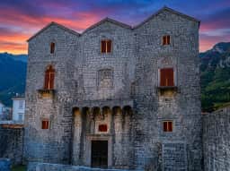 The roofless stone shell of the Tre Sorelle palace on the Prčanj shore, its Gothic facade pierced by three tall pointed windows.