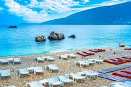 Playa de guijarros con hileras de tumbonas blancas, rocas en agua turquesa cristalina y un cabo verde cerca de Herceg Novi