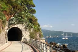Paseo marítimo que entra en un túnel de piedra junto a rocas y barandillas, con veleros en la bahía de Herceg Novi