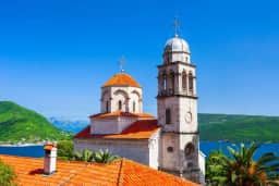 Iglesia ortodoxa con cúpula de tejas naranjas y campanario de piedra sobre tejados rojos y la bahía en Herceg Novi