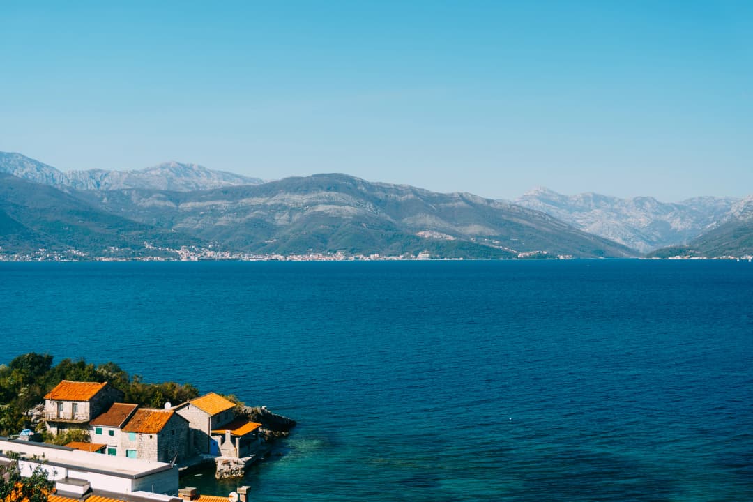 Wide view across the Bay of Kotor from the Luštica side, with waterfront houses below and mountains opposite