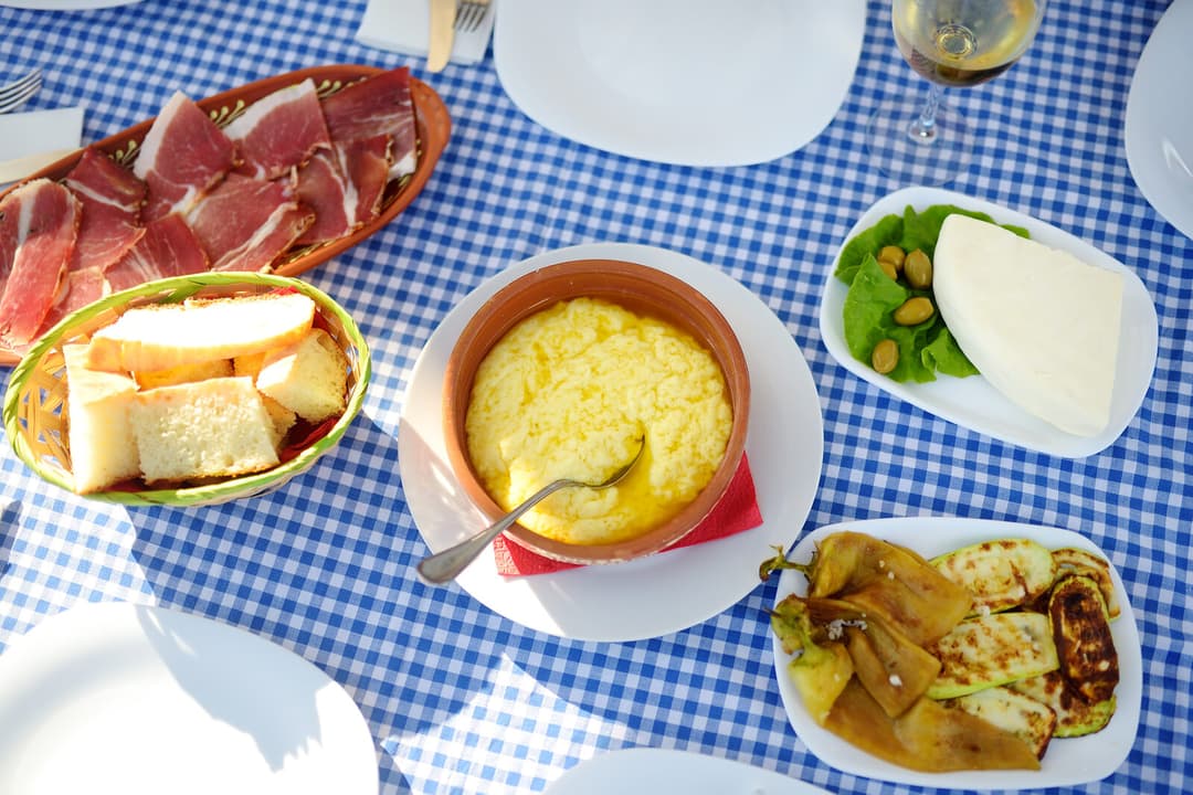 Traditional kacamak served in a clay pot on a blue checkered tablecloth with prsut and cheese