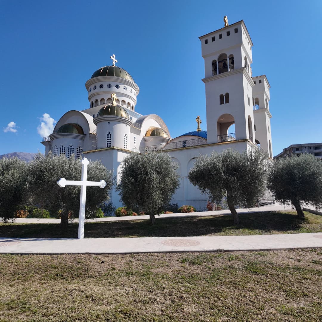 Bijeli krst među starim maslinama na tratini pred hramom, a iza krošanja se dižu zvonici i pozlaćena kupola