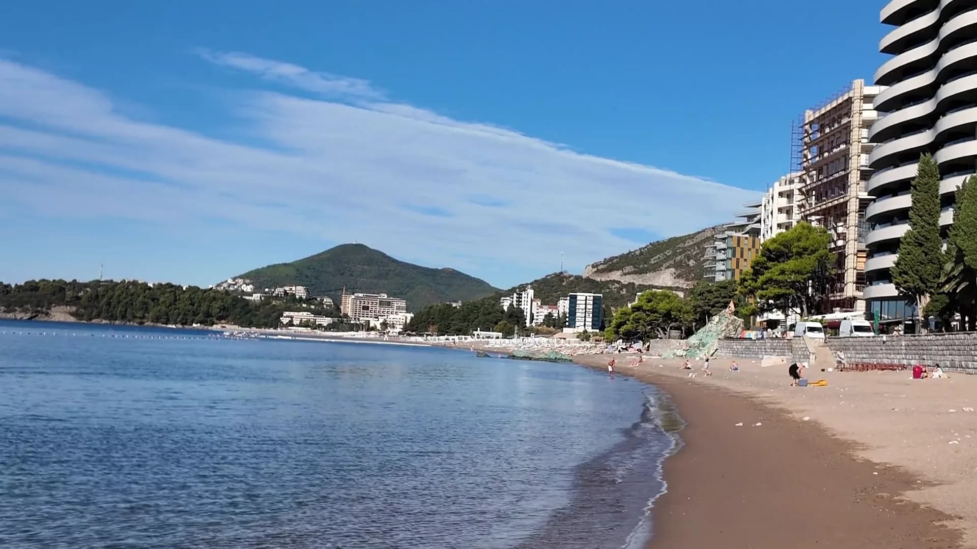 The curve of Bečići beach on a September morning, the shallows running out to the pine-covered headland and the town of Rafailovići across the bay
