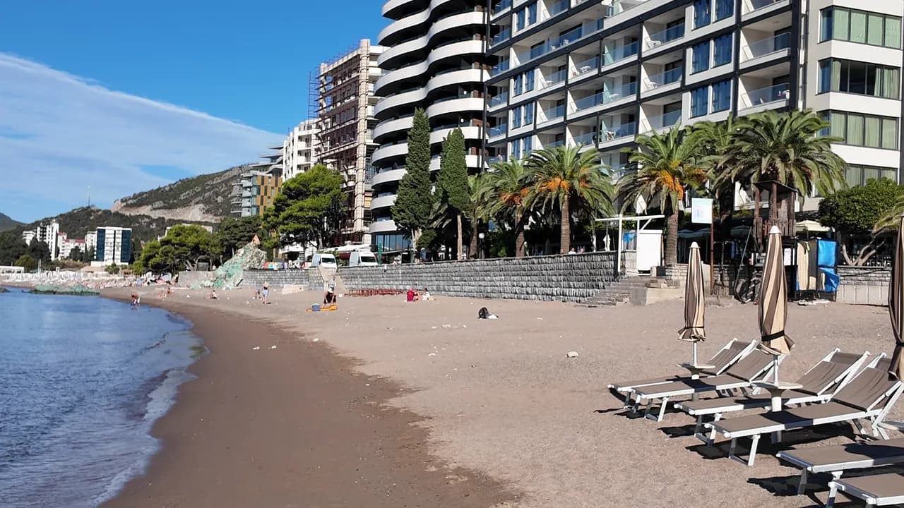 Bečići beach on a September morning: rows of umbrellas on the dark sand, walkway mats down to the water and the pine-covered headland closing the bay