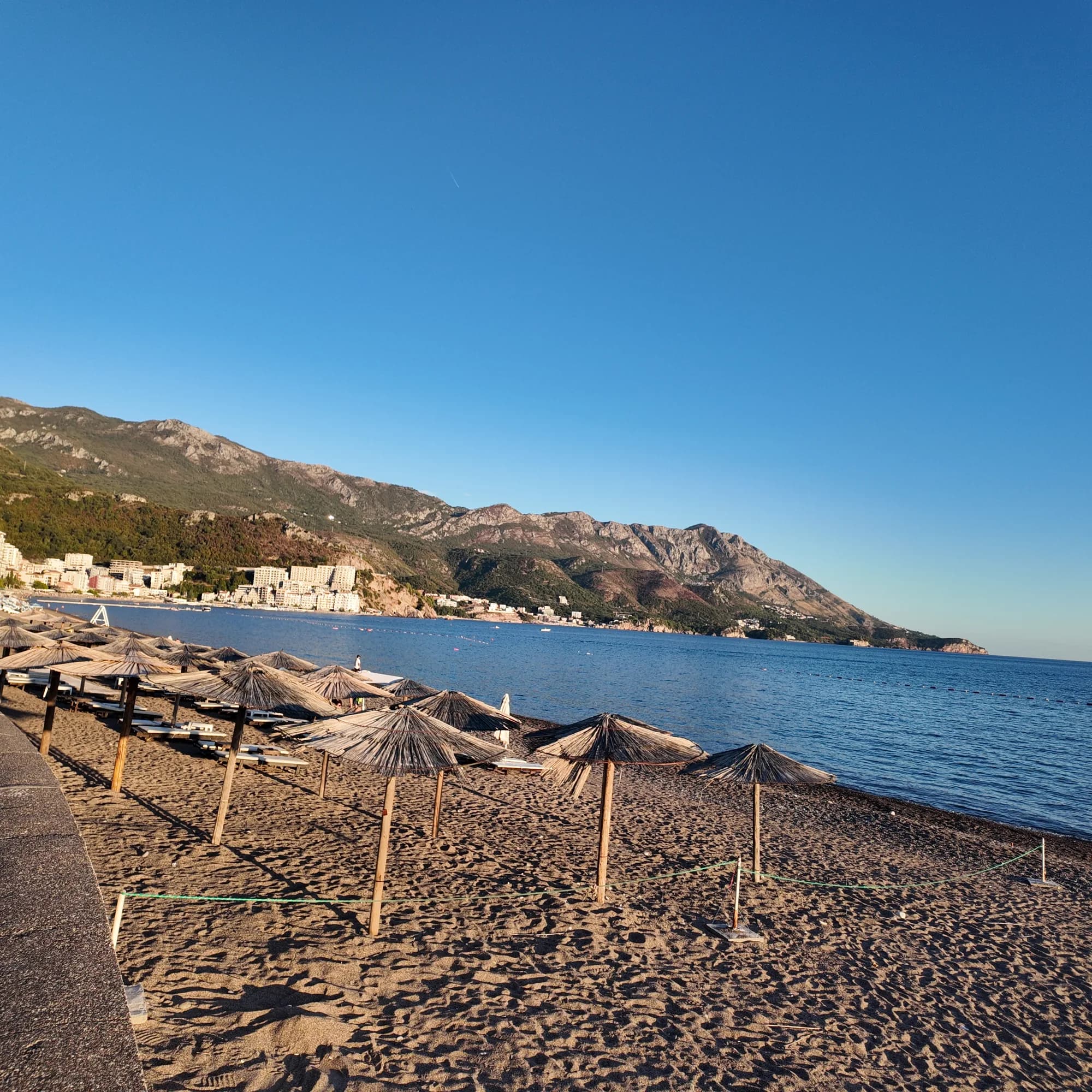 Roped-off sectors of umbrellas at the eastern end of Bečići in evening light, Rafailovići and its headland behind