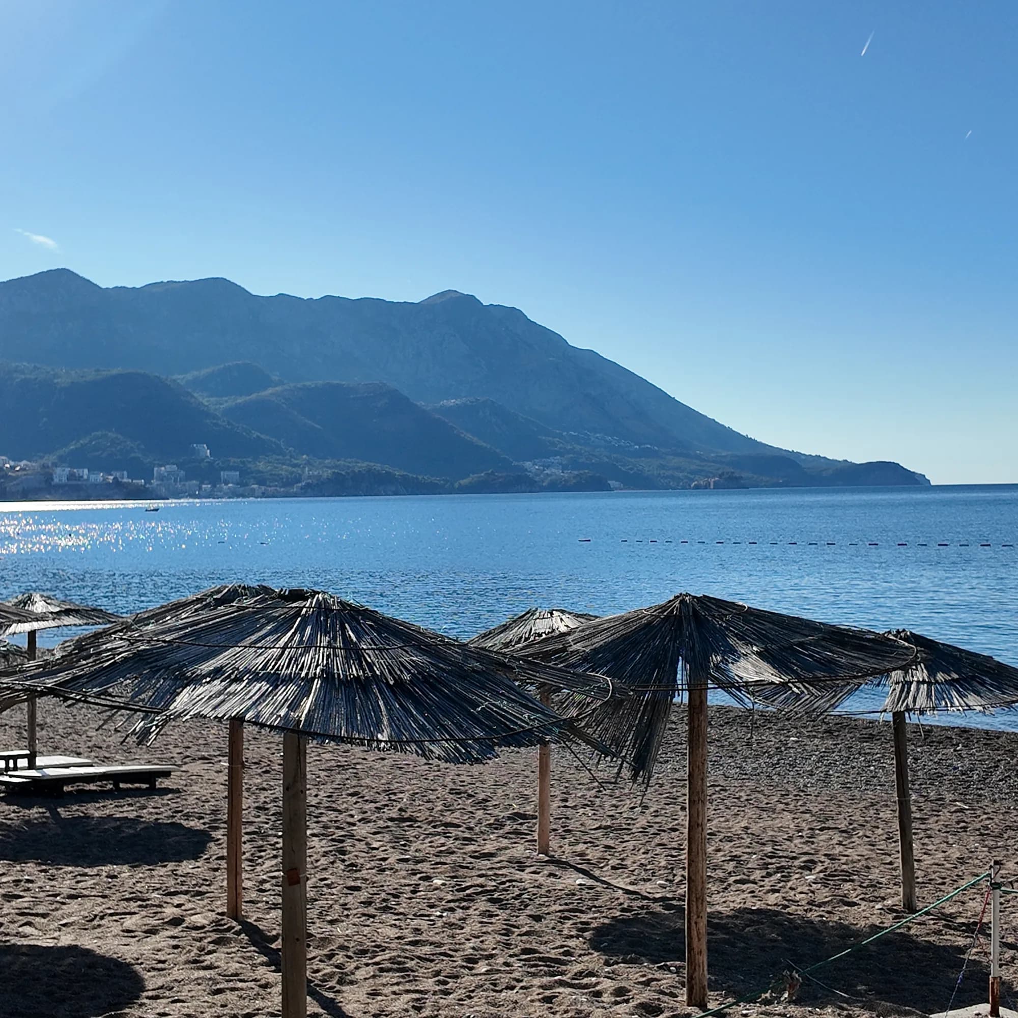 Thatched umbrellas on the dark sand of Bečići in the morning, the buildings of Rafailovići and the ridge across the bay