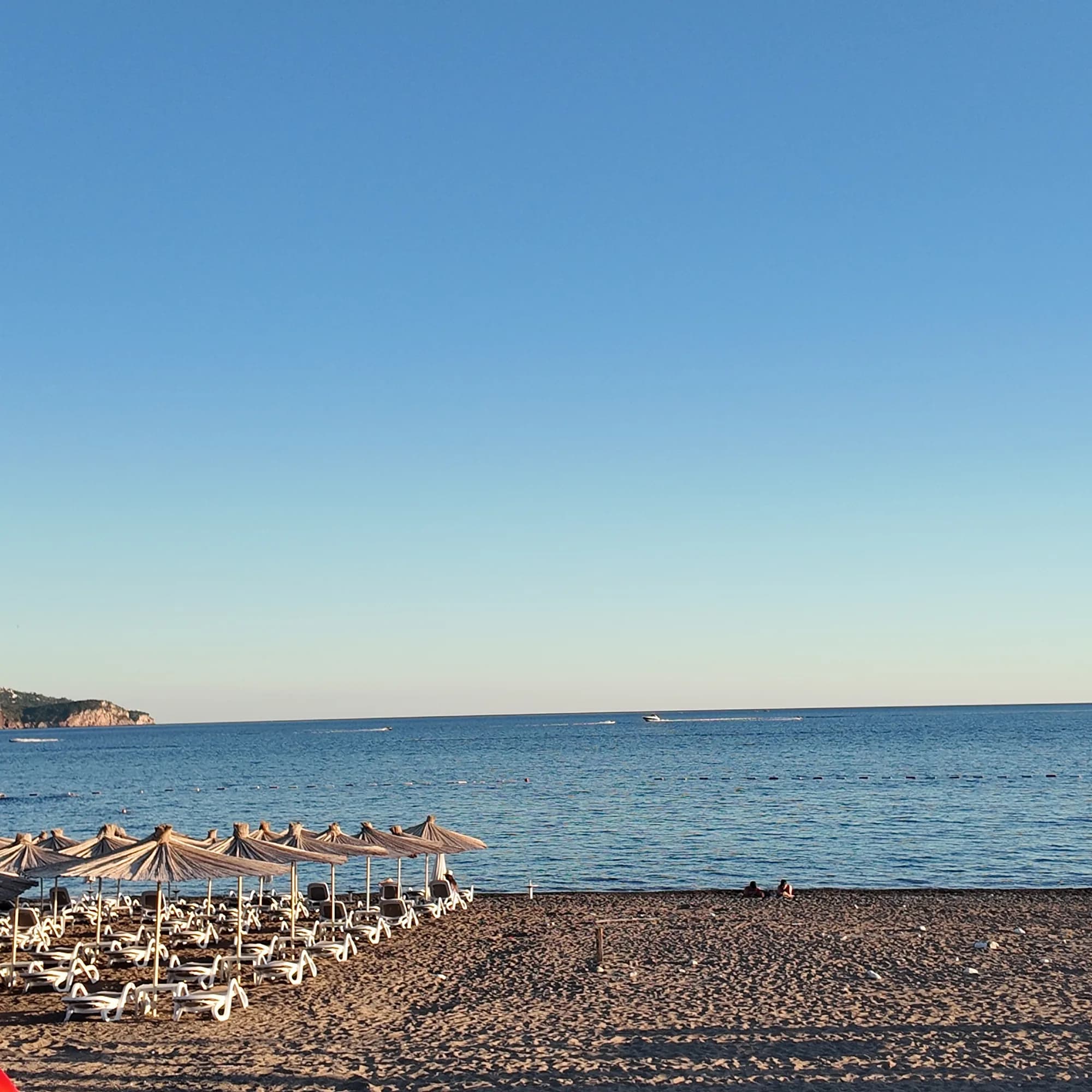 Thatched umbrellas and white loungers at the water’s edge at Bečići, boats in the bay and the Sveti Stefan headland to the east