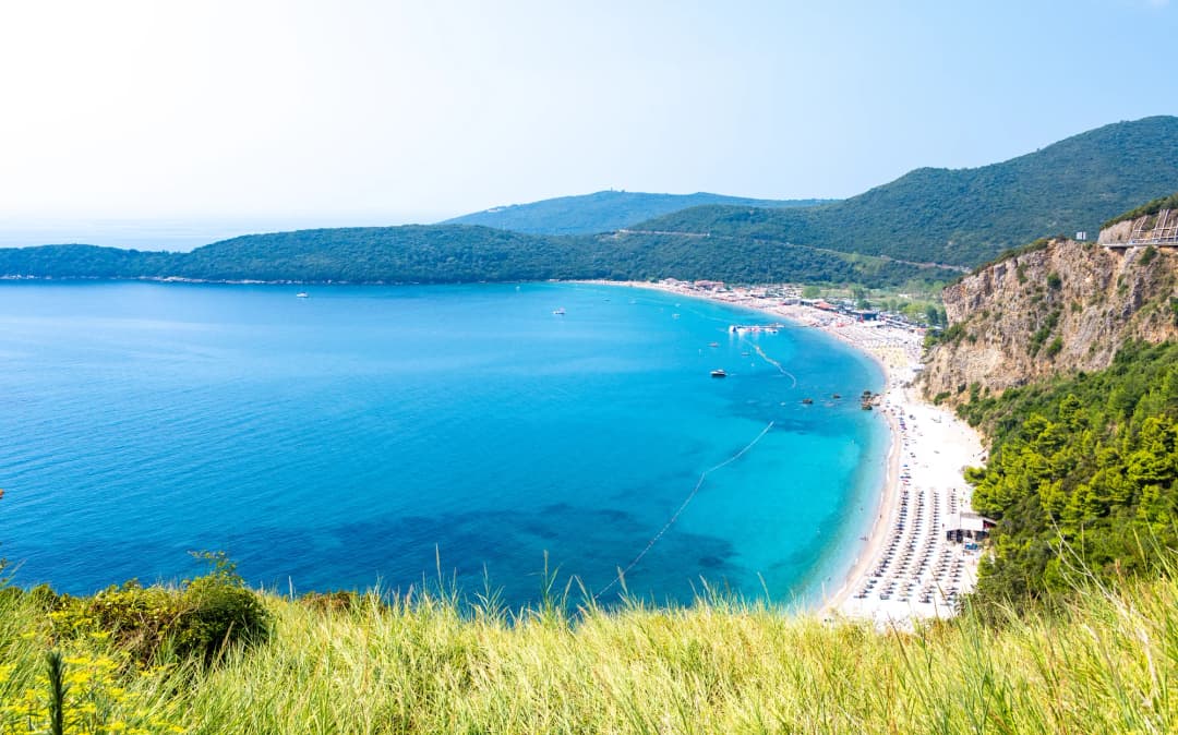 Posjetioci šetaju obalskom stazom kroz park Miločer između Svetog Stefana i plaže Miločer, sa mediteranskim borovima koji uokviruju pogled na more