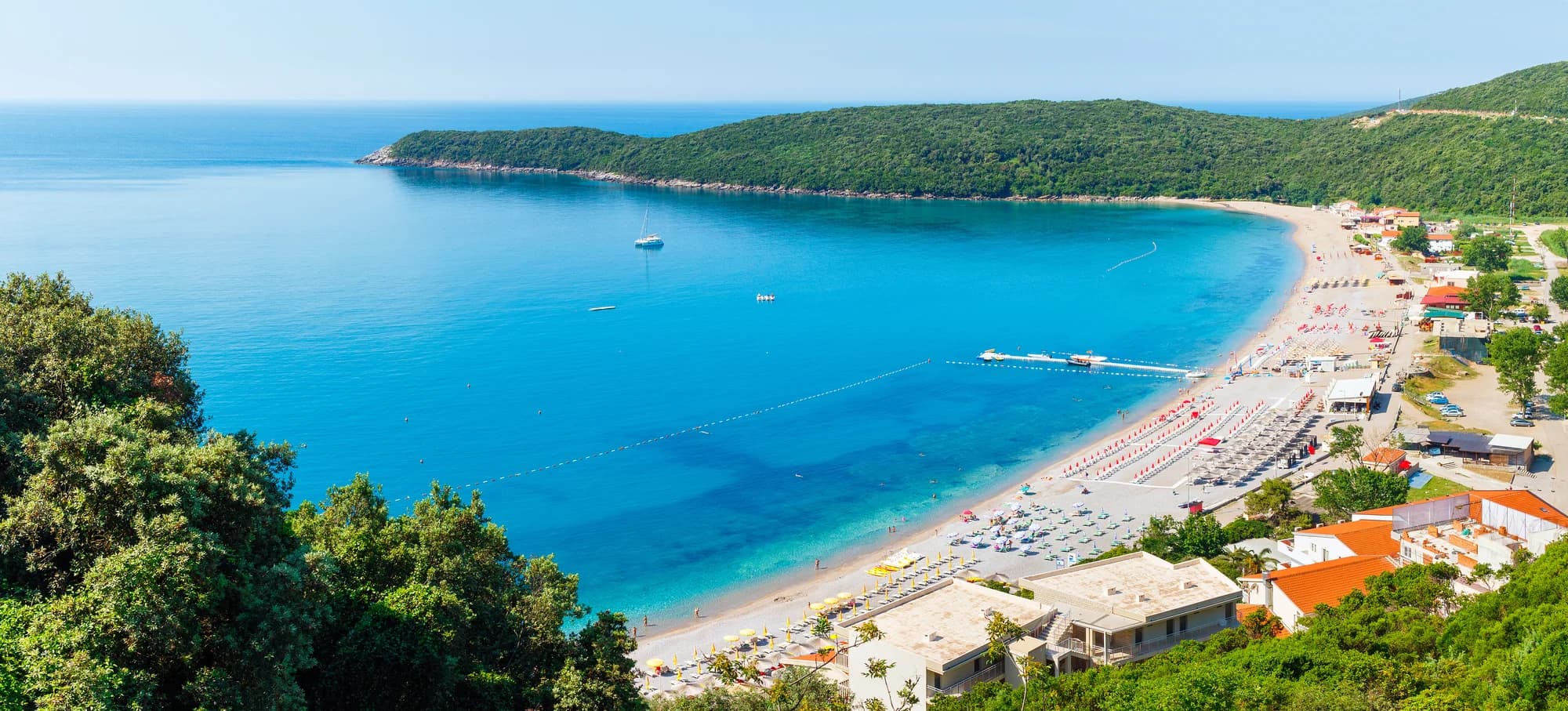 The organised western section of Jaz from the hill: rows of umbrellas, a jetty, the houses and car parks behind and the green headland across the bay