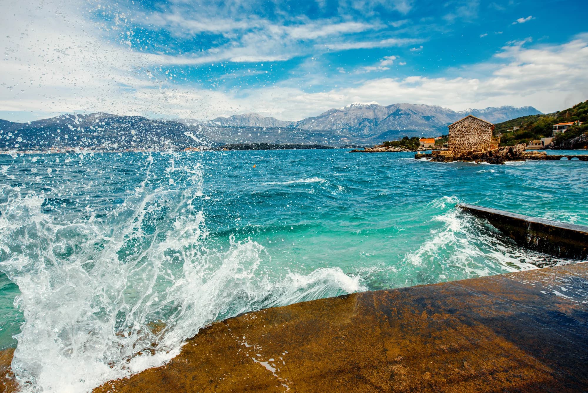A wave breaking over the quay at Bjelila, the stone house on the point and snow on the mountains across the bay