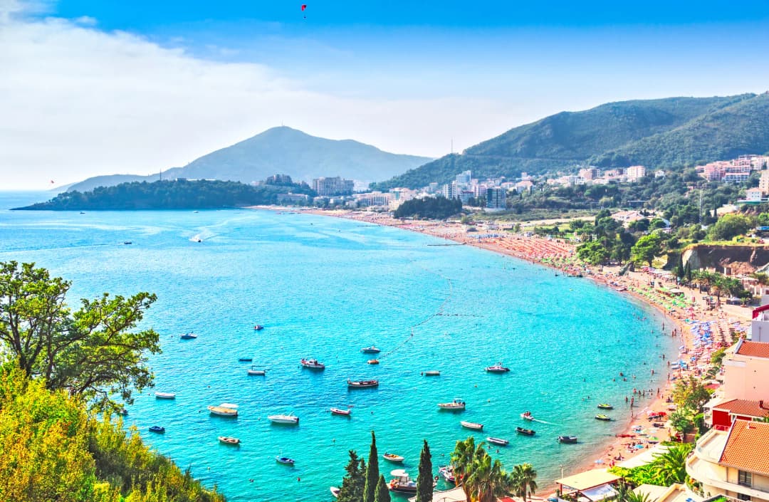 Bečići and Rafailovići from the hillside above, a long curve of sand with boats moored offshore and rows of umbrellas running the length of the shore.