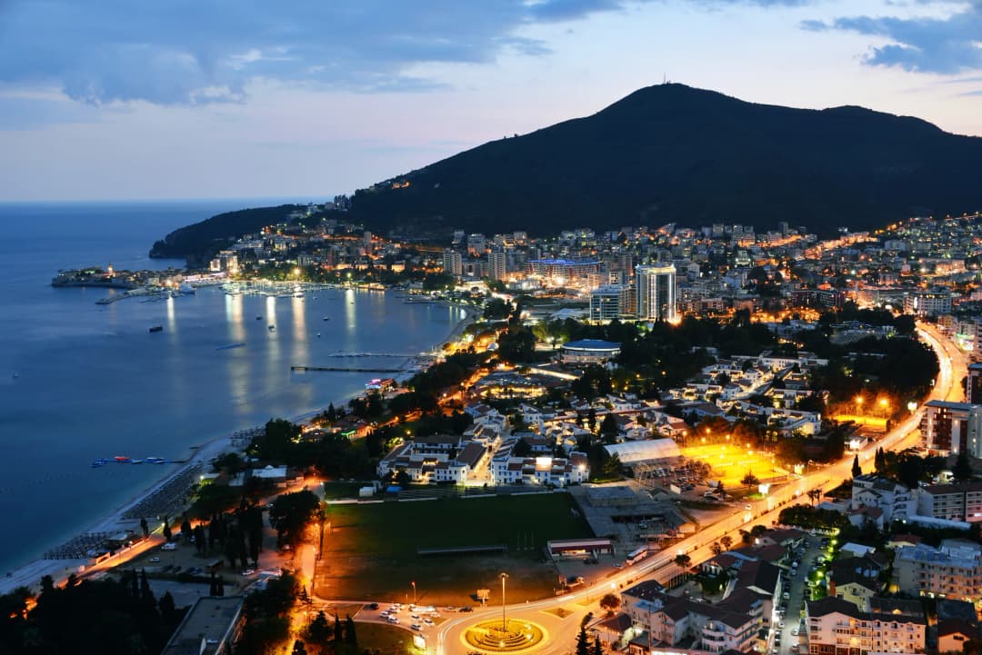 Budva lit up after dark, seen from the hill above: the peninsula, the bay and the coast road picked out in lights.