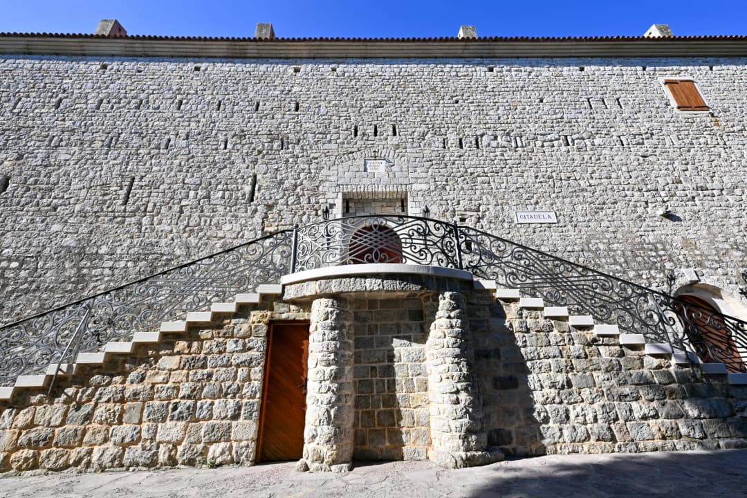 The Citadela entrance in Budva Old Town: twin curved staircases with wrought-iron balustrades rising to a landing above the arched doorway, under the fortress wall.