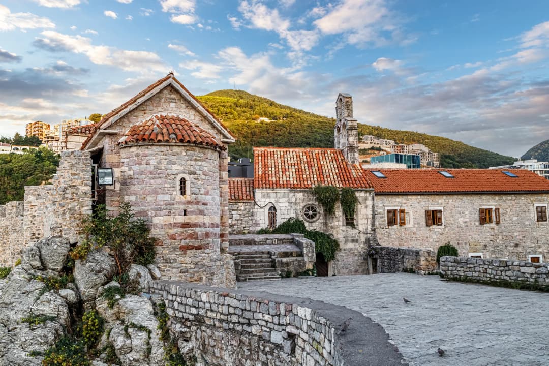 Stone church roofs and a round bell tower inside Budva Old Town, with the citadel wall rising behind them.
