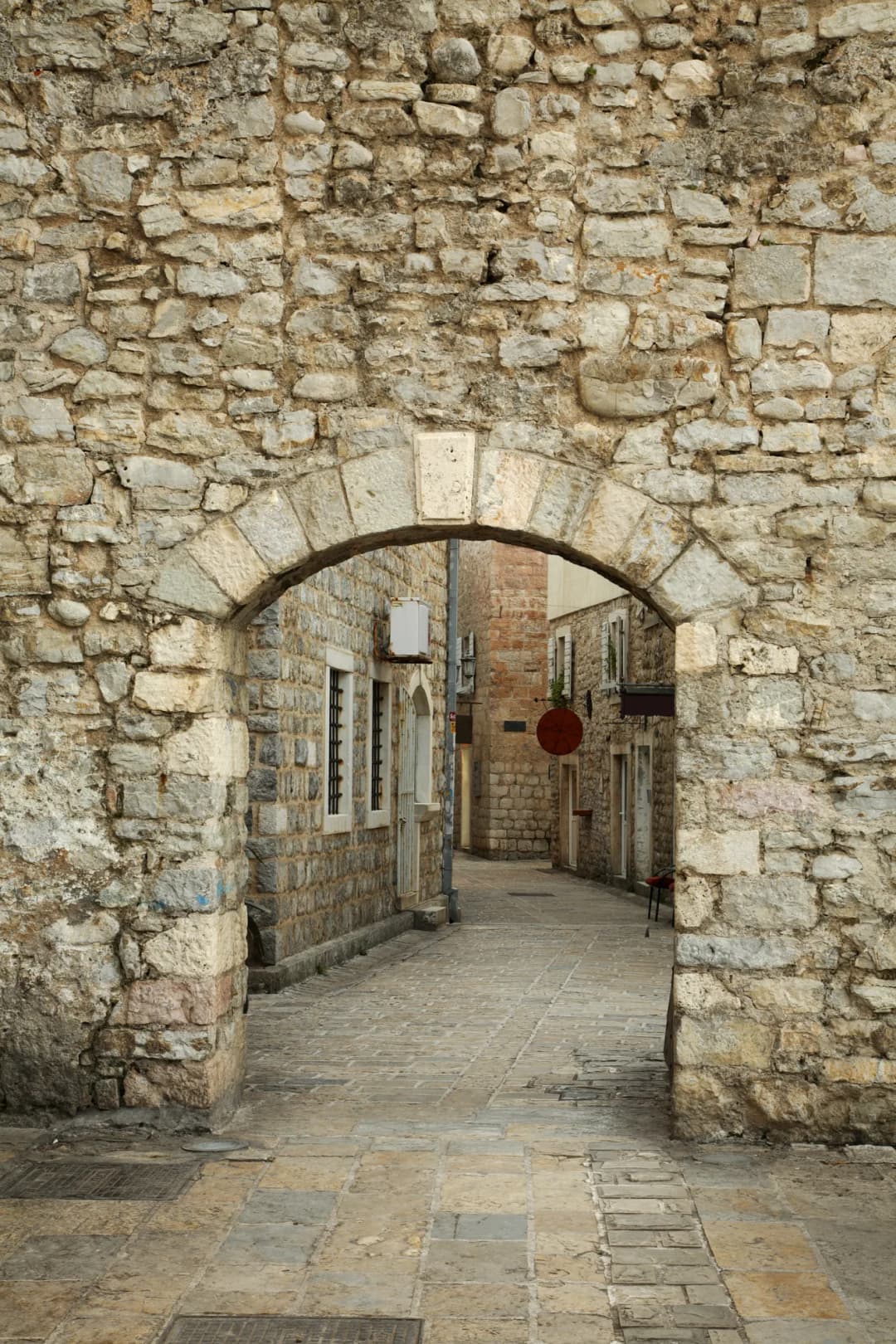 A stone gate arch cut through Budva Old Town's wall, opening onto a paved lane of shuttered stone houses.