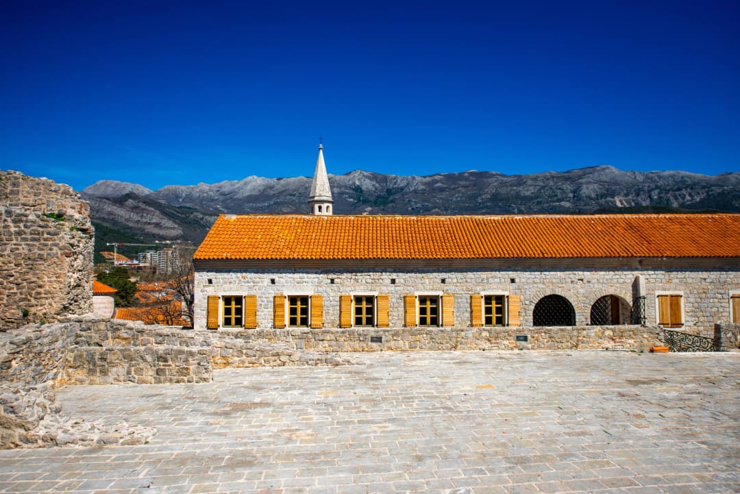 A long arcaded stone building under new terracotta tile inside Budva Old Town, a bell tower behind it.