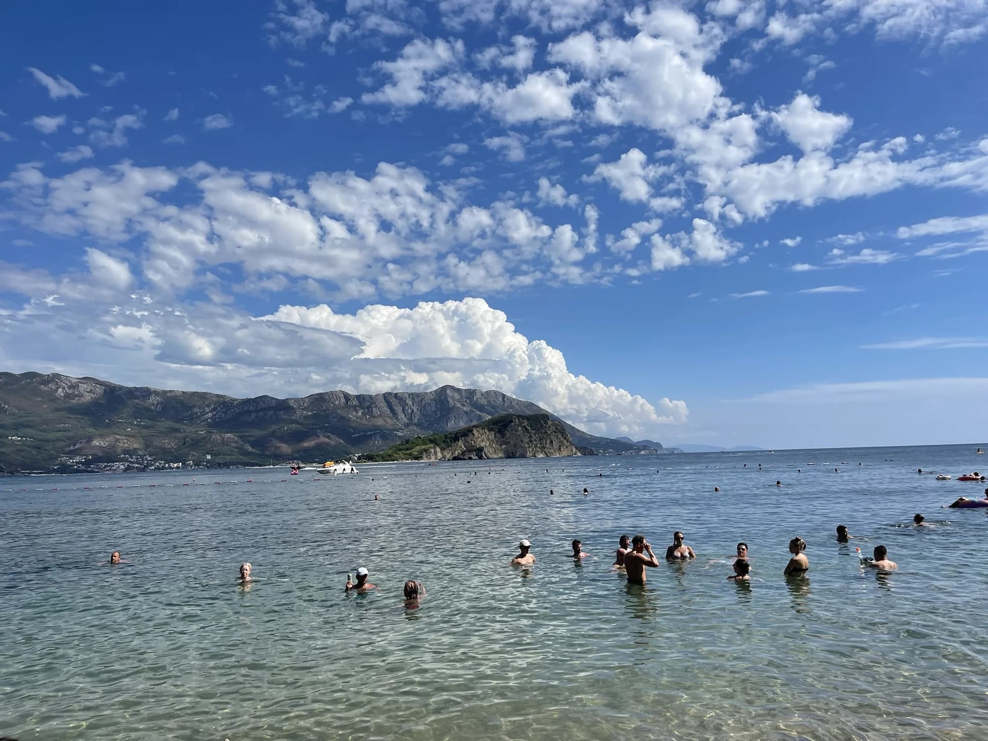 Mogren II from the sand: swimmers standing in the shallows, the wooded point of Sveti Nikola island and the mountains behind Budva