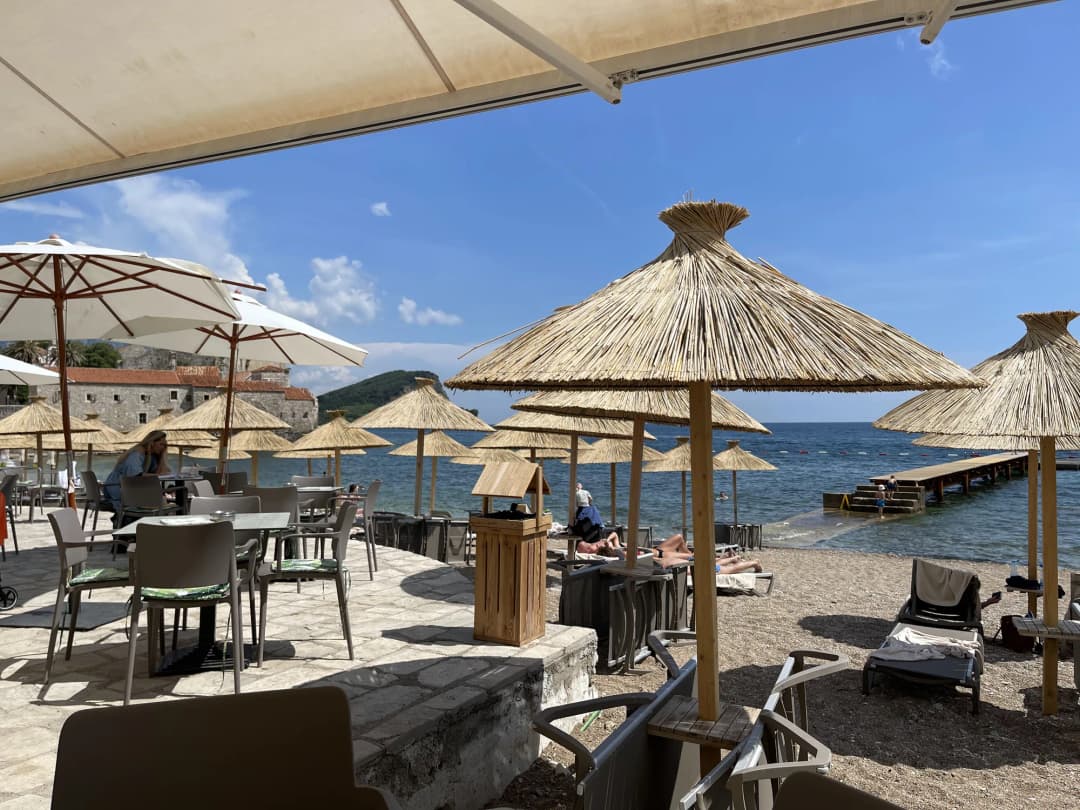 Ričardova Glava beach: thatched parasols and sunbeds on a short strip of sand, a beach-bar terrace behind and the Old Town wall closing the left of the bay.