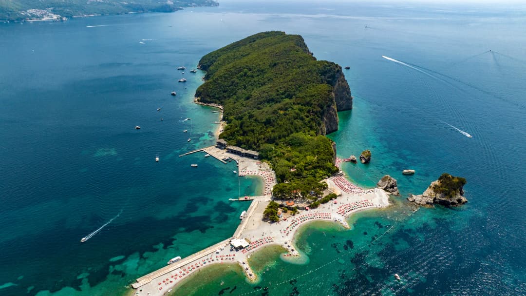 Sveti Nikola island from the air, its wooded ridge rising above the sandbar beaches and their rows of umbrellas, with Budva on the shore beyond.