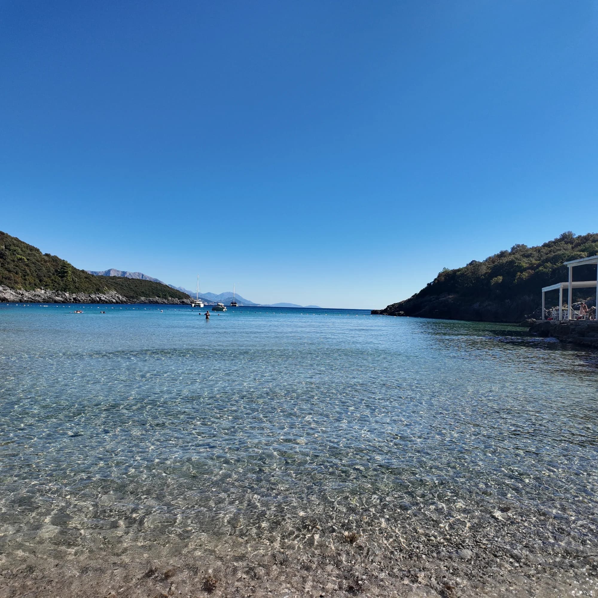 The shallow cove at Trsteno from the water’s edge: a bather standing far out, the buoy line, anchored yachts and the beach-club pergola on the rocks