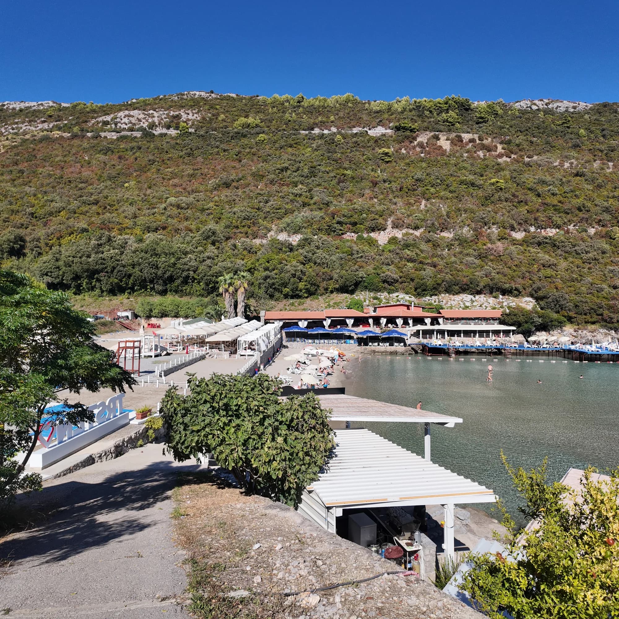 The beach club at Trsteno from the track above: white umbrellas and cabanas on the sand, the restaurant terraces and the Trsteno sign