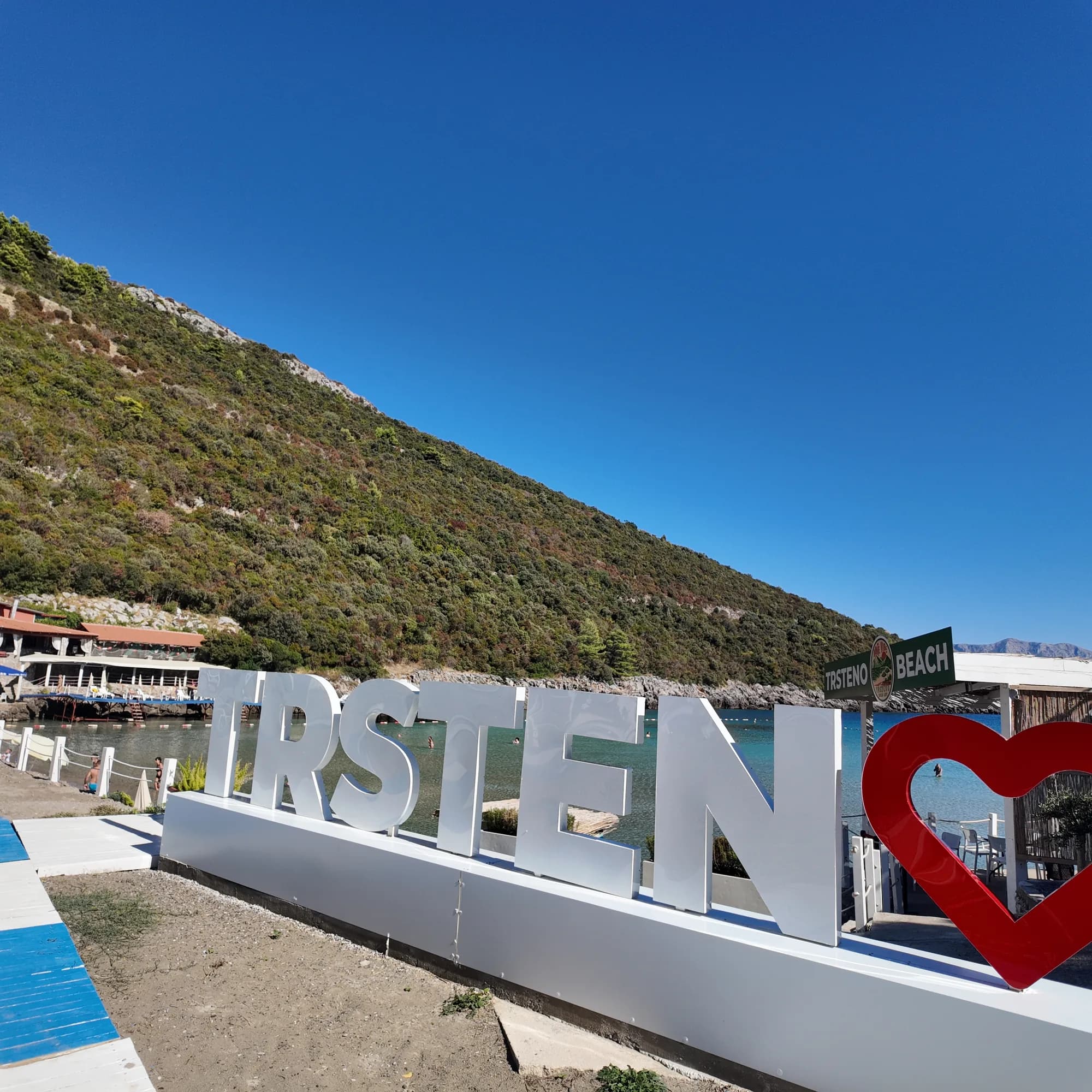 The white TRSTENO letters and red heart at the top of the beach, the Trsteno Beach sign and the restaurant behind