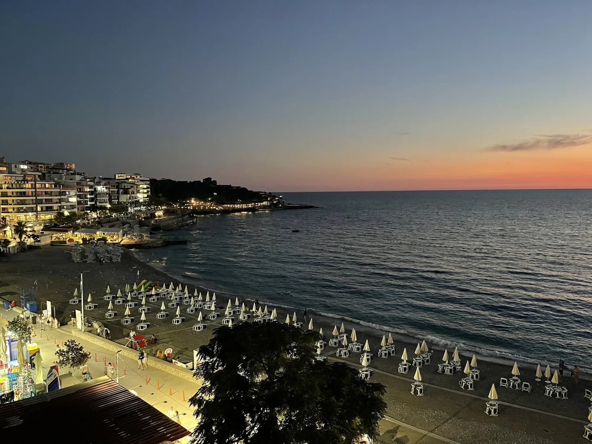 The Dobra Voda waterfront at dusk: lit apartment blocks, the promenade and folded umbrellas along the beach