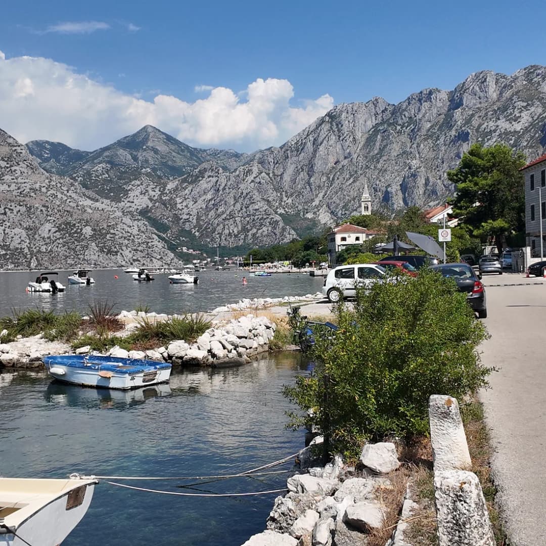 Kamene kuće duž obale Boke Kotorske, s niskim kamenim molom i malim čamcima privezanim u mirnoj vodi pod planinama.