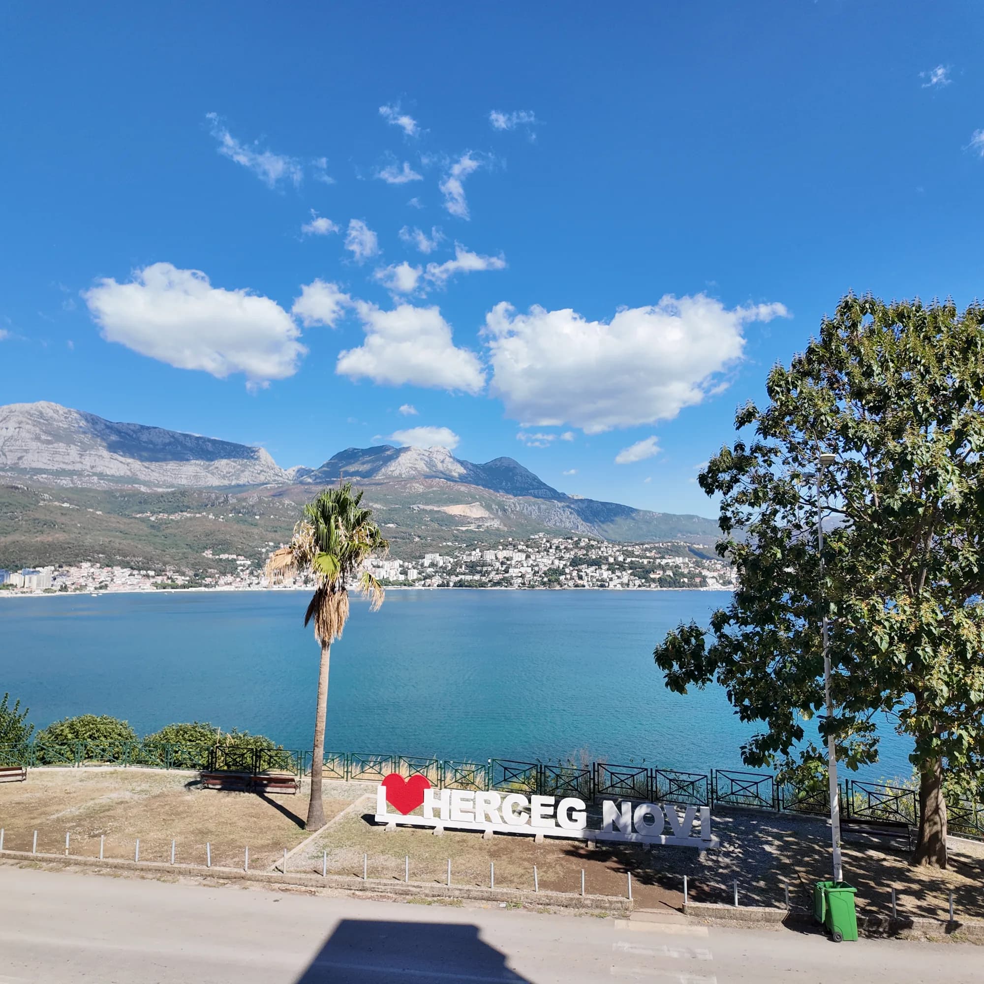 Il cartello I ♥ Herceg Novi sulla strada di Njivice, con la città distesa lungo la sponda opposta alle sue spalle