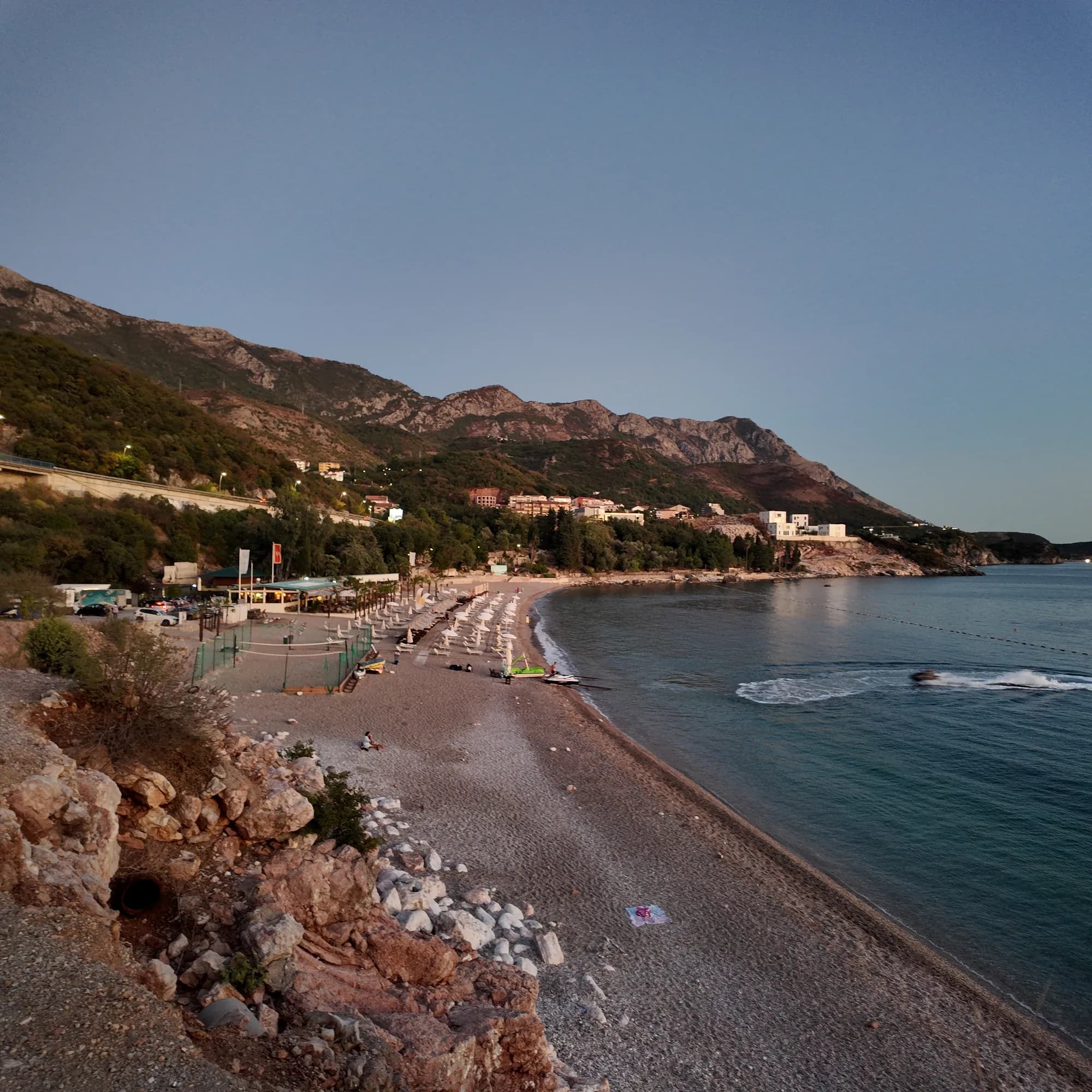 Kamenovo beach and its bay under the bare hills in evening light, sunbeds on the pebbles and a boat lane marked by buoys