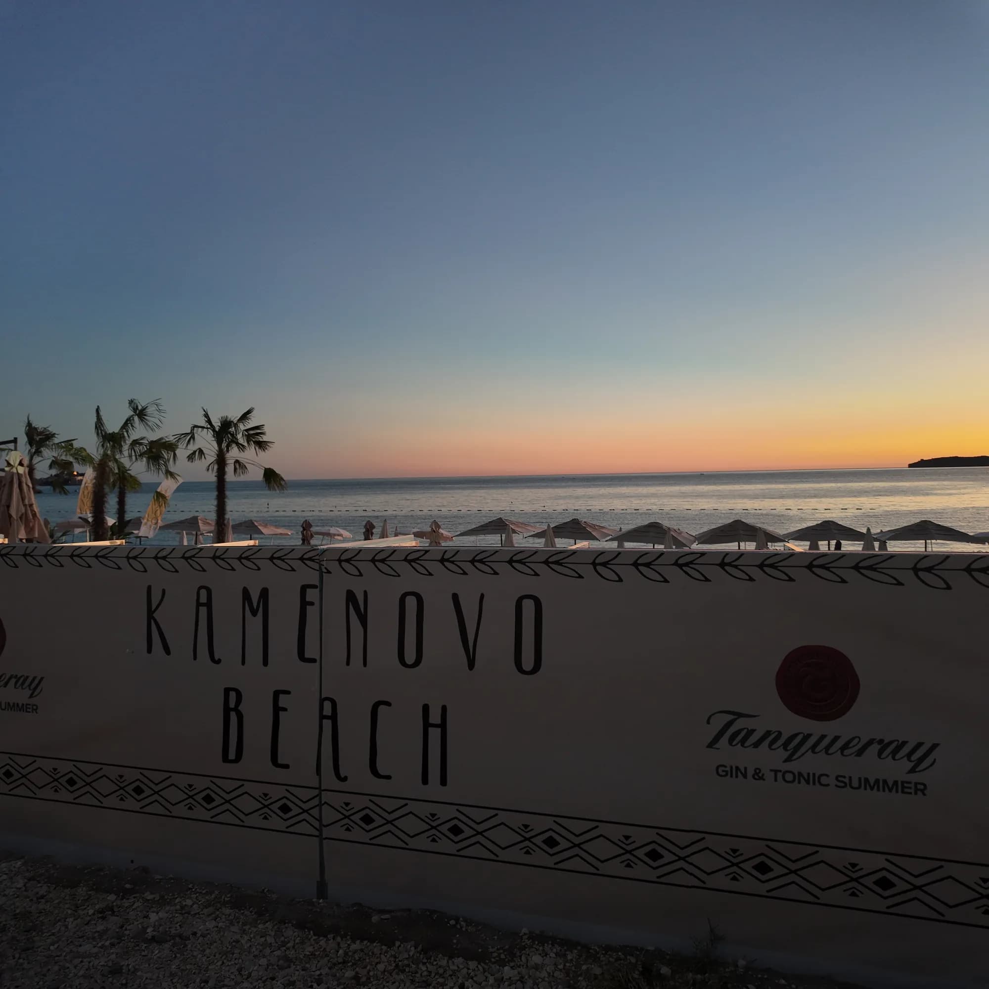 The Kamenovo Beach sign at the entrance to the beach bar, palms and umbrellas behind it at dusk