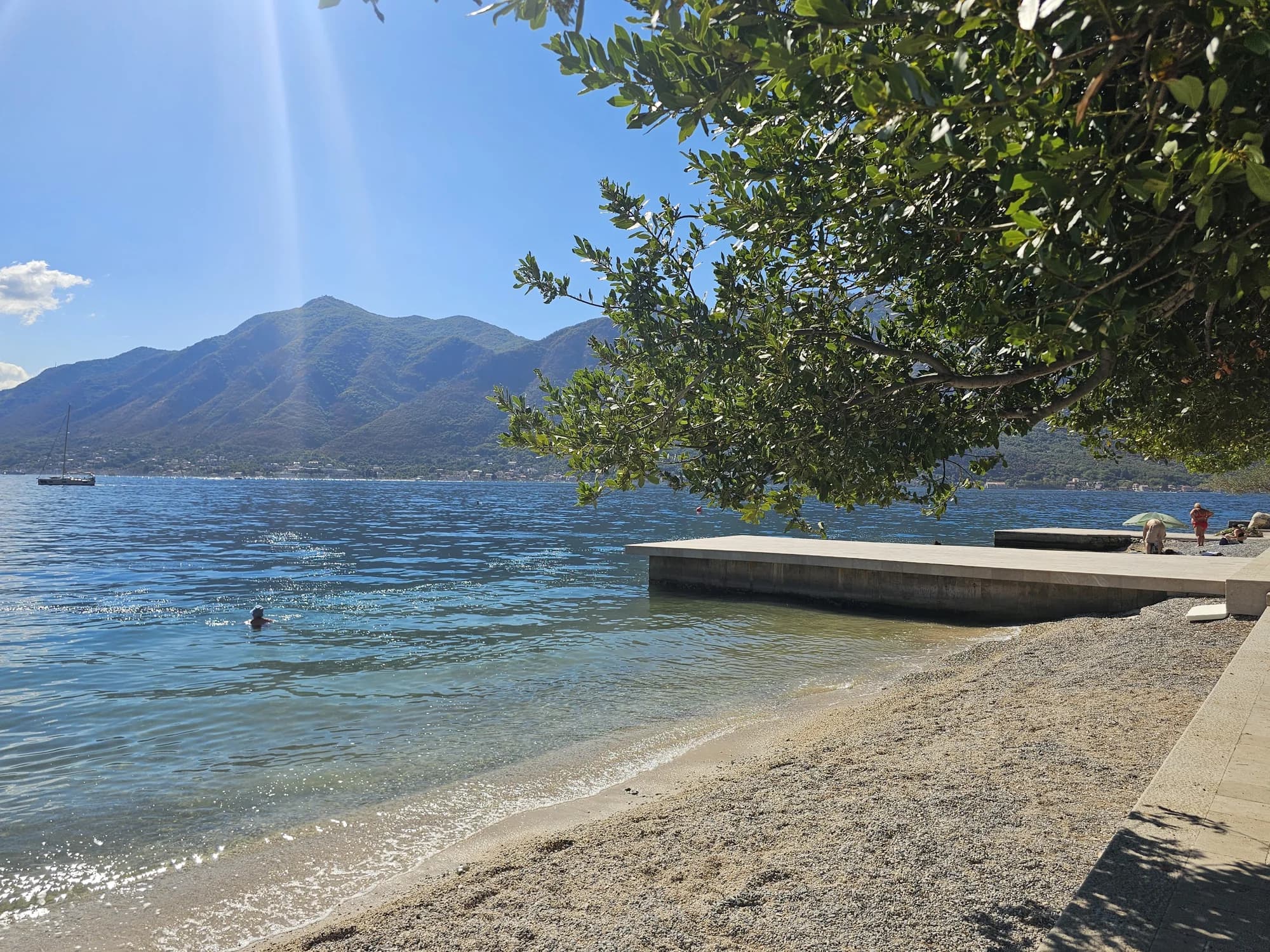 The clear shallows, pebble shore and swimming platform at Bajova Kula framed by leafy shade