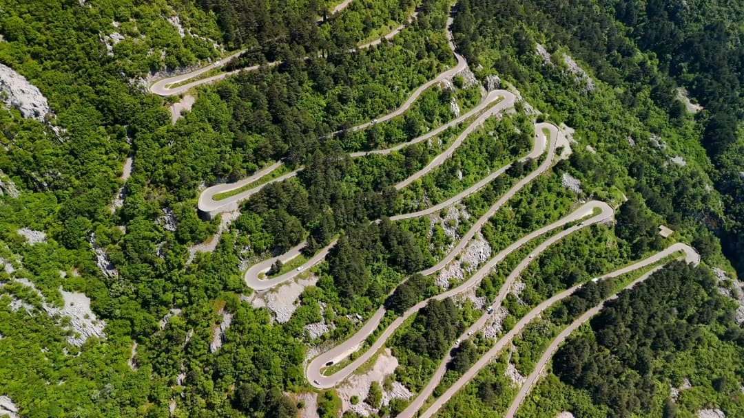 The same serpentine hairpins from the air in 2026, the road threading through dense green forest with cars on the bends