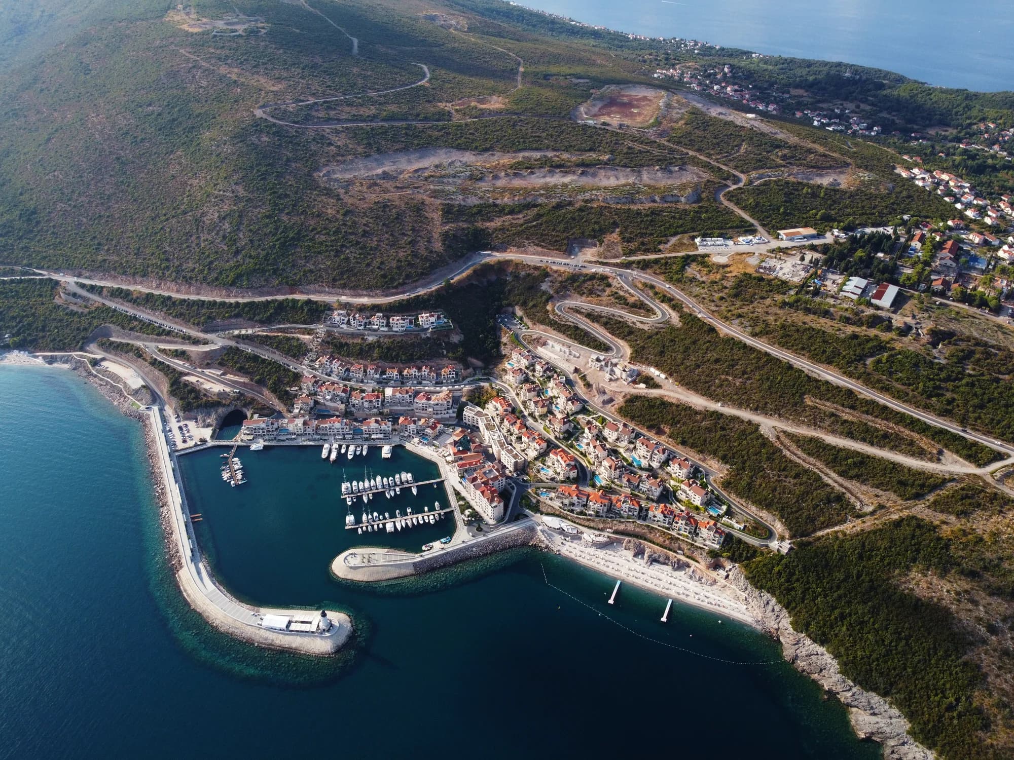 The southern coast of Luštica from the air at Luštica Bay, the marina village and its breakwater — the same shore a few kilometres east of Plavi Horizonti