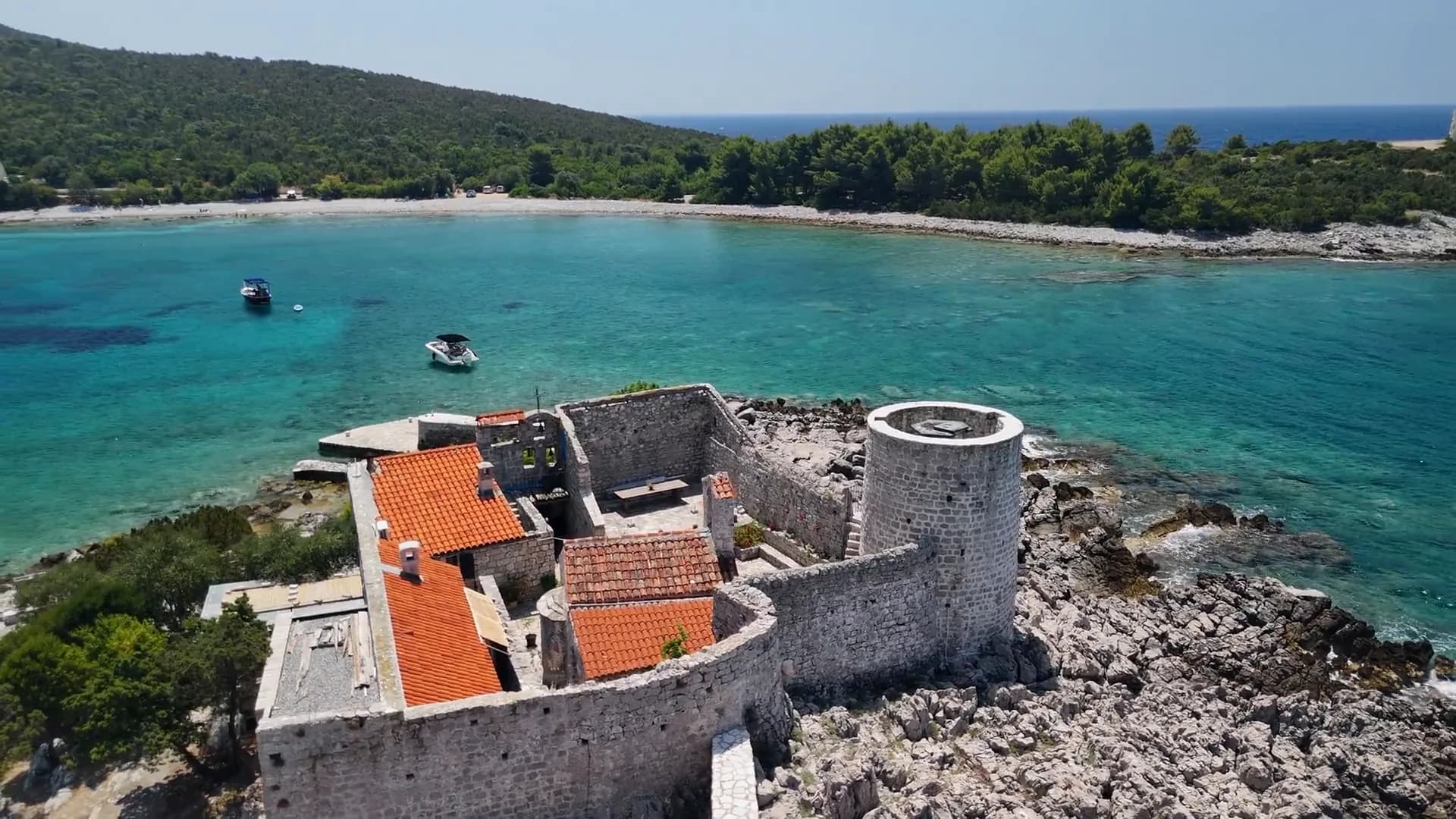 The Arza fort on its rocky point at the tip of Luštica, and behind it the bay of Mirišta: boats at anchor over turquoise shallows and the pale shore of the beach