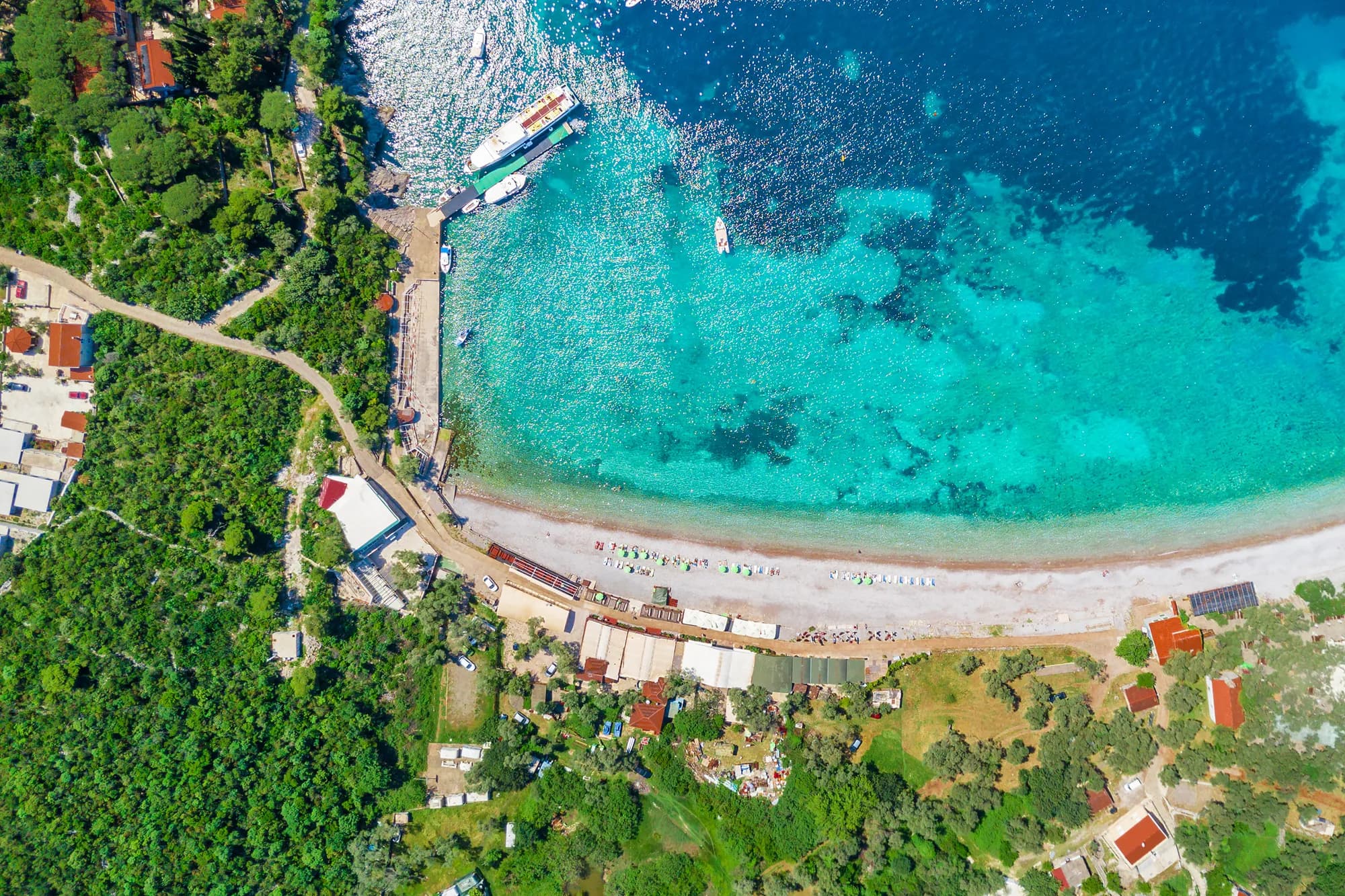 Žanjice direktno odozgo: plaža i njen mol, izletnički čamac pored njega i put koji se spušta kroz masline