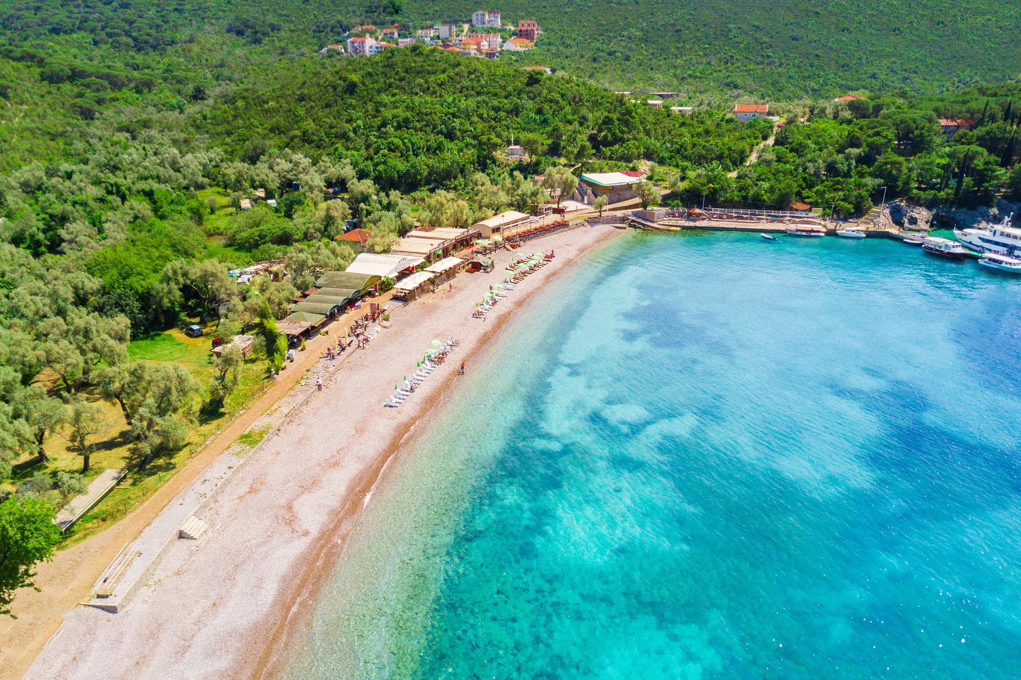 The pebble arc of Žanjice from the air, loungers under the olives along the back of the beach and the water shading from clear shallows to deep turquoise
