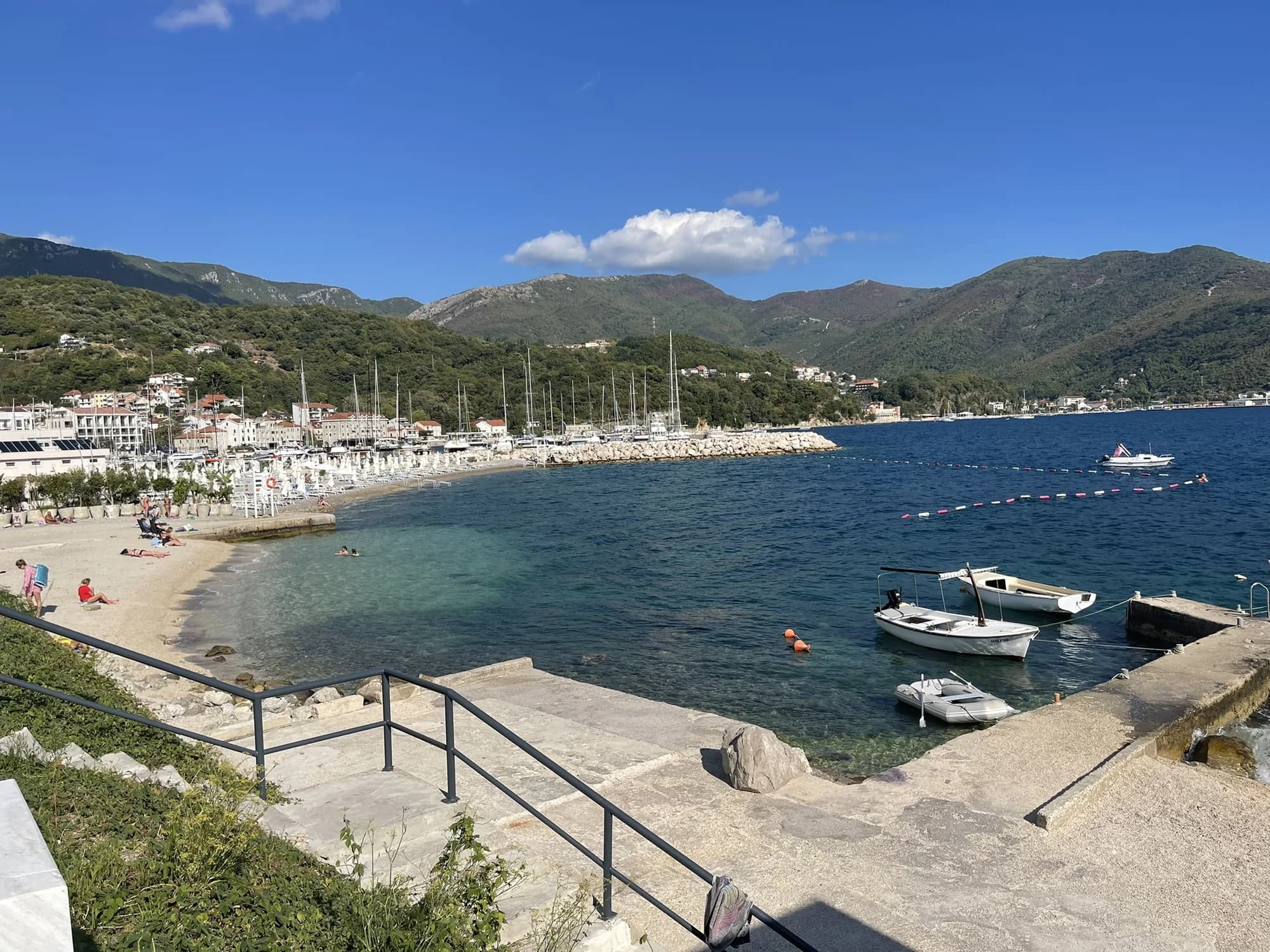 Meljine’s public pebble beach and clear buoyed water beside Lazure marina, with the green mountains behind