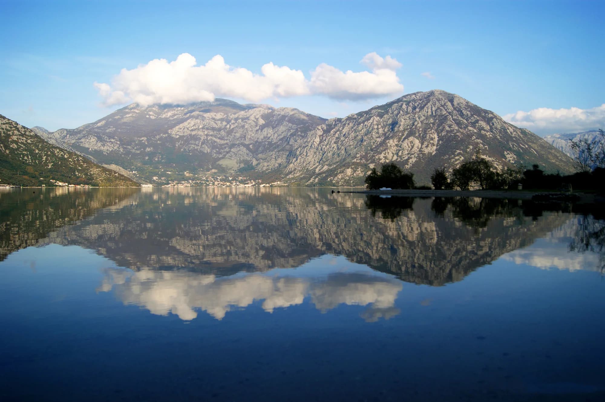 The Risan basin from the Morinj shore on a flat day, the mountains and clouds reflected and a pebble spit with trees at the right