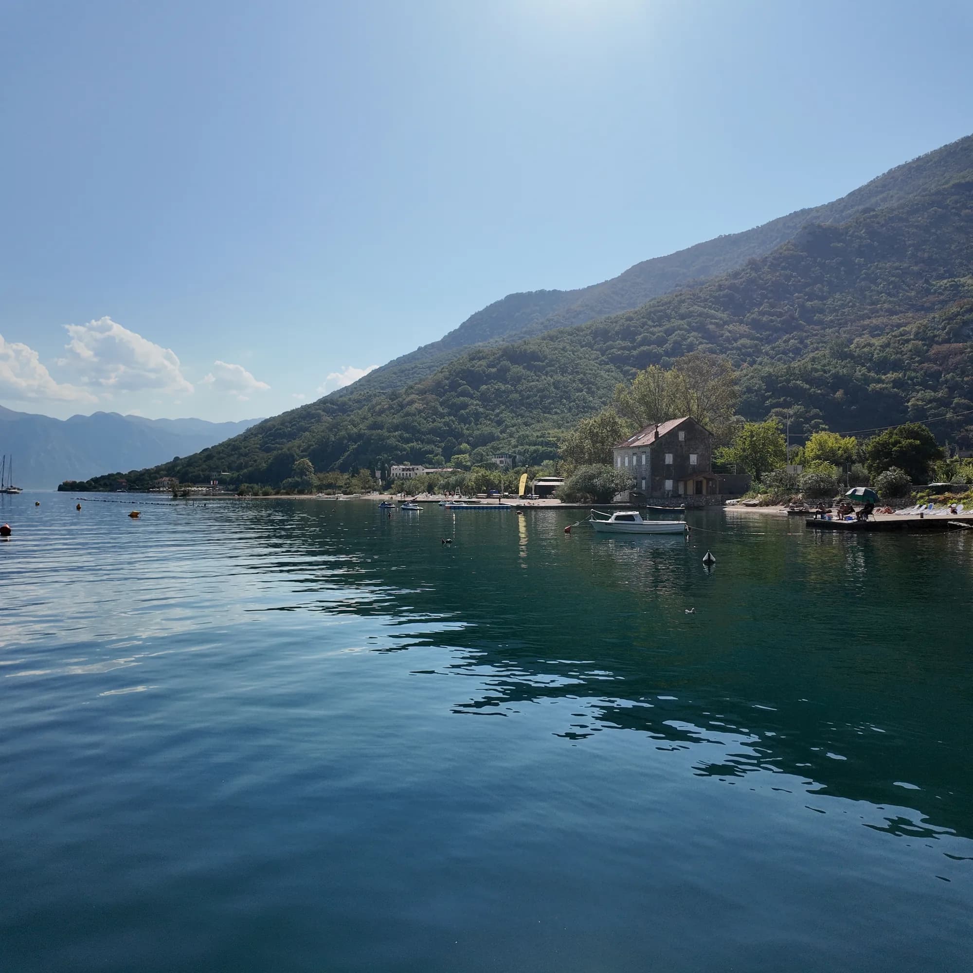 Morinj beach from the water: loungers on the pale shore, pedal boats at the buoy line, the stone house at the stream mouth and the wooded hill above