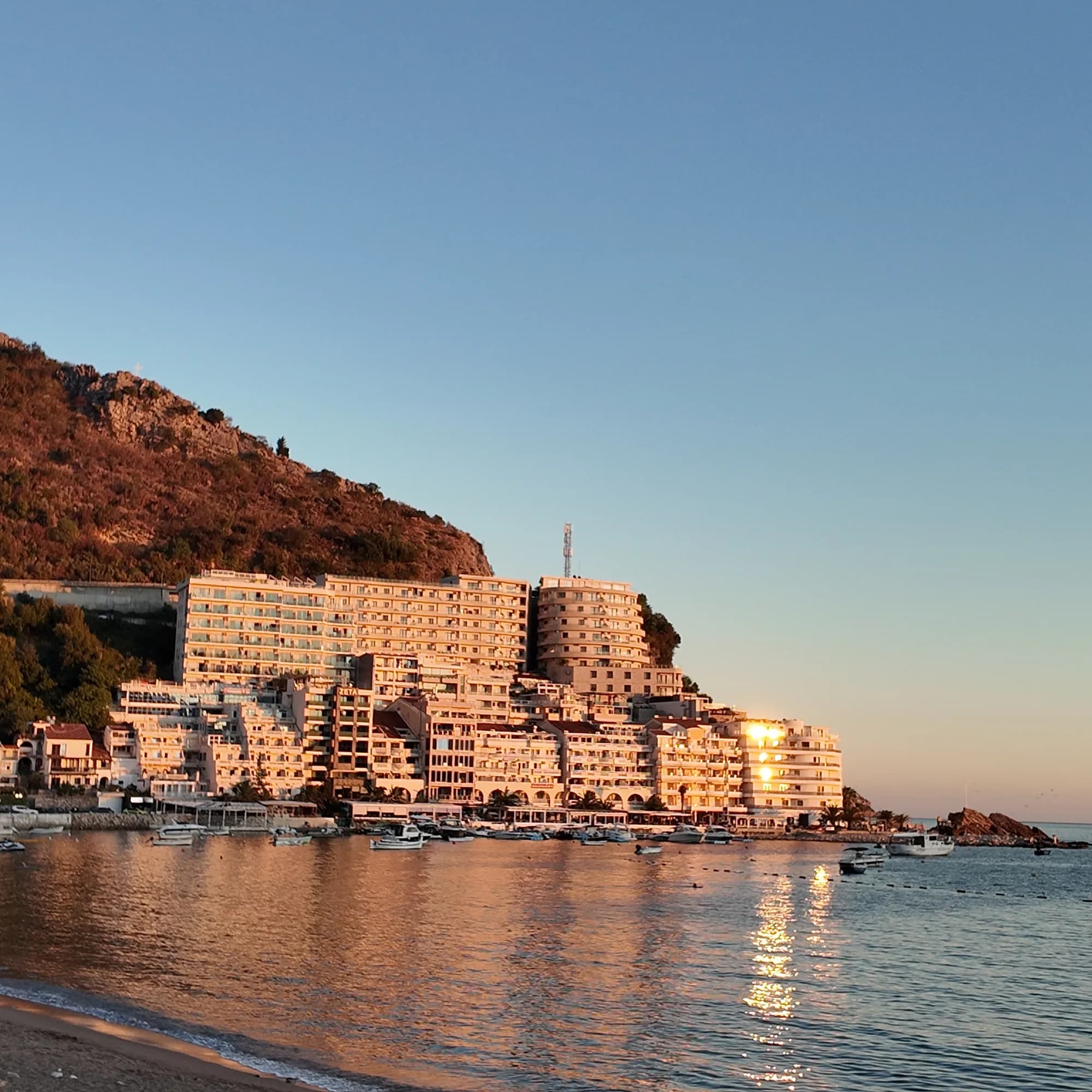 The terraced apartment block on the headland at Rafailovići lit orange by the setting sun, the pebble beach and calm sea in front of it