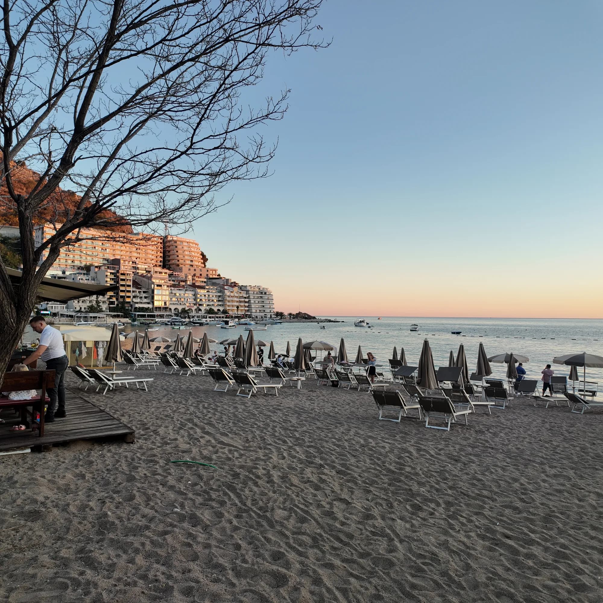 Grey umbrellas and loungers on the sand at dusk, a waiter laying tables under a tamarisk, the headland blocks lit pink