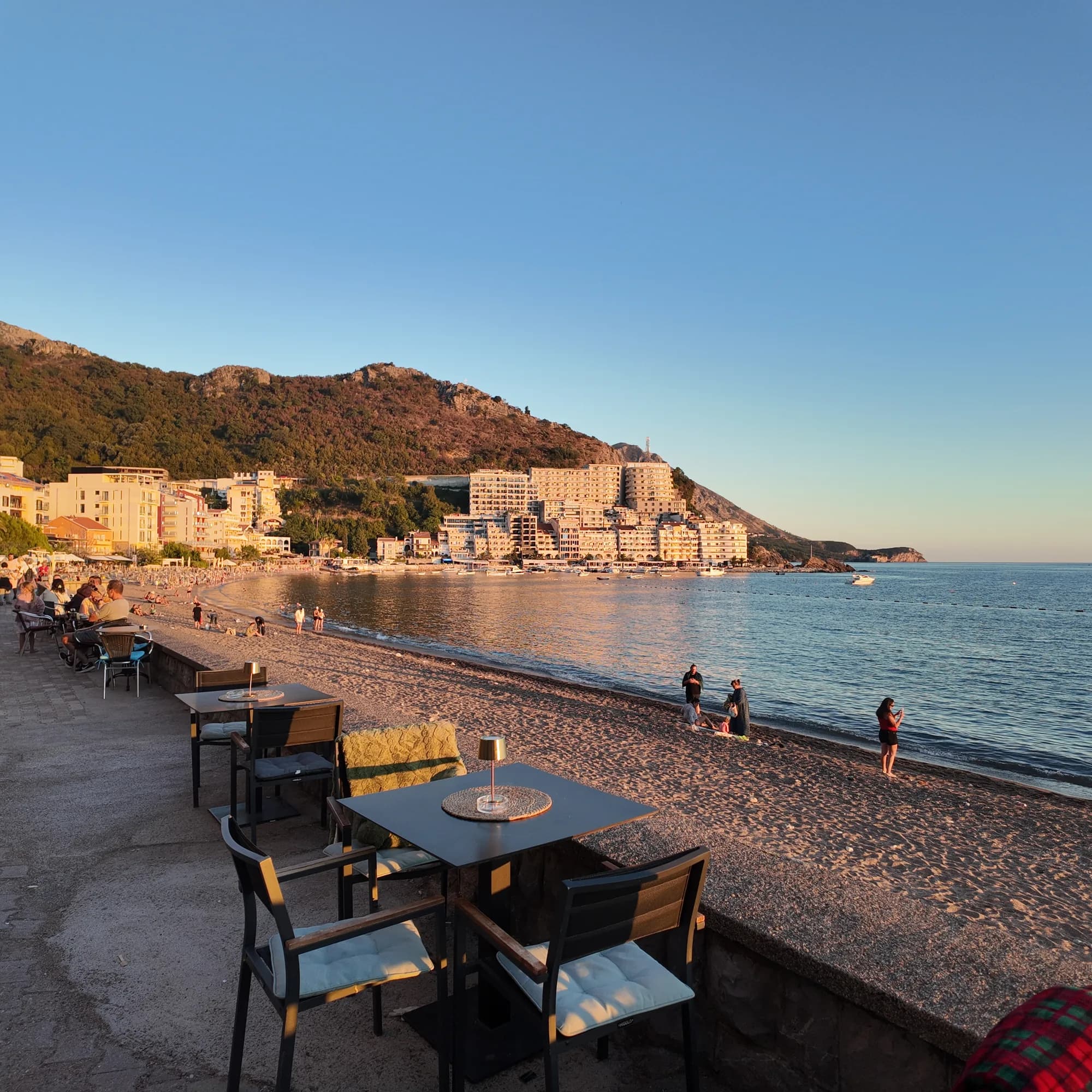 Restaurant tables on the promenade wall at sunset, the sand-and-pebble beach curving to the village and the headland