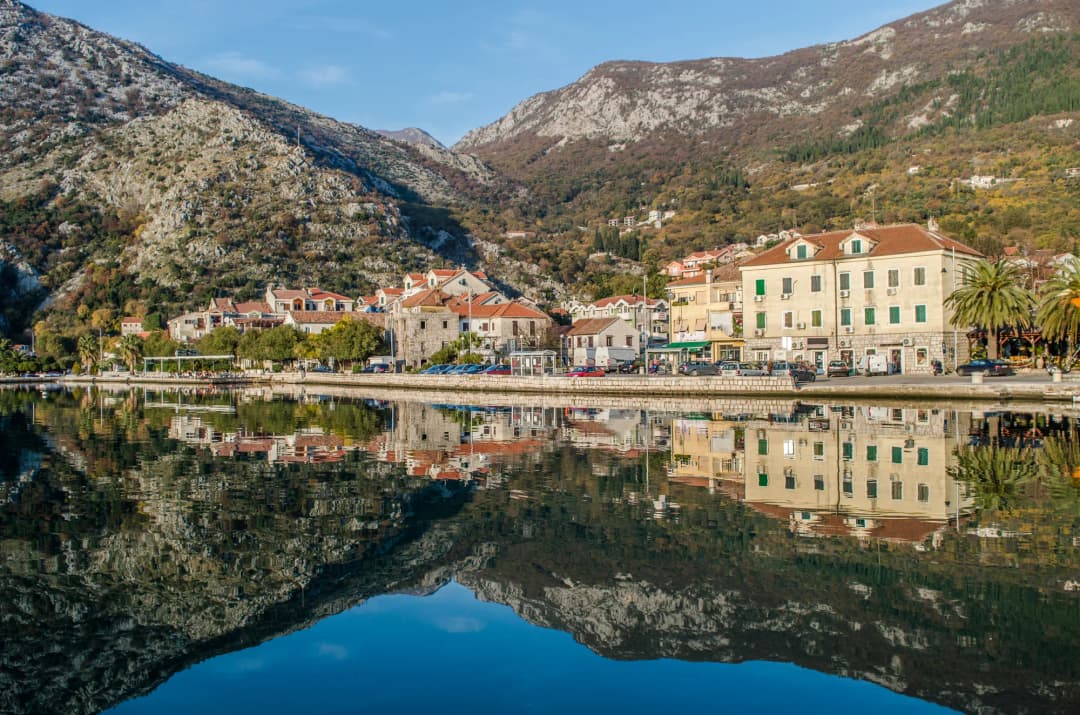 The Risan waterfront reflected in still water, with the bay's mountains rising behind the town's stone houses and palms.